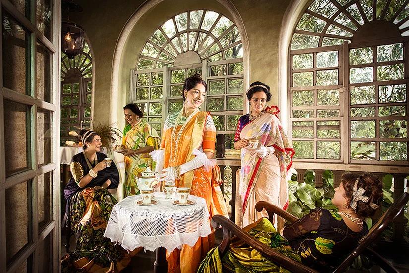 Women in saris gathered around a lace-covered tea table in a heritage room with arched windows